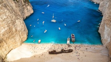 İyon Denizi kıyısındaki Navagio sahilinin insansız hava aracı görüntüsü. Paslı bir geminin yanında dinlenen insanlar, mavi su.