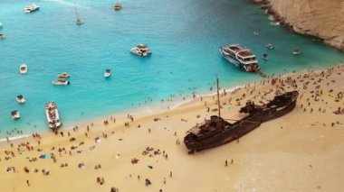 Yunanistan 'ın Zakynthos kentindeki İyon Denizi kıyısındaki Navagio plajının insansız hava aracı görüntüsü. Demir atmış tekneler, dinlenen insanlar, paslı gemi, mavi su. Yavaş çekim