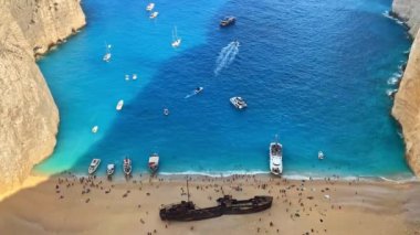 Yunanistan 'ın Zakynthos kentindeki İyon Denizi kıyısındaki Navagio plajının insansız hava aracı görüntüsü. Demir atmış tekneler, dinlenen insanlar, paslı gemi, mavi su. Yavaş çekim