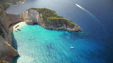 Yunanistan 'ın Zakynthos kentindeki İyon Denizi kıyısındaki Navagio plajının insansız hava aracı görüntüsü. Kayalıklar, demirli ve yüzen tekneler, dinlenen insanlar, mavi su.