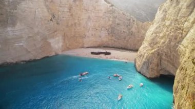 Yunanistan 'ın Zakynthos kentindeki İyon Denizi kıyısındaki Navagio plajının insansız hava aracı görüntüsü. Kayalıklar, demirli ve yüzen tekneler, dinlenen insanlar, mavi su.