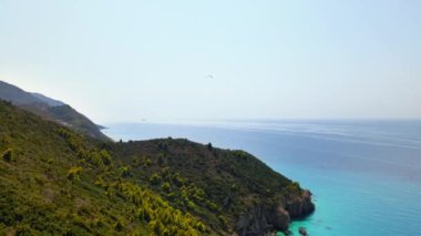 Yunanistan 'ın Zakynthos kentindeki İyon Denizi kıyılarının insansız hava aracı görüntüsü. Tepe yeşillikle kaplıydı, adam paraşütle süzülüyordu, mavi su.