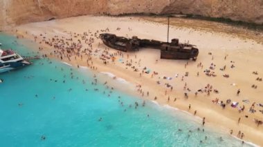 Yunanistan 'ın Zakynthos kentindeki İyon Denizi kıyısındaki Navagio plajının insansız hava aracı görüntüsü. Demir atmış tekneler, dinlenen insanlar, paslı gemi, mavi su.