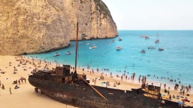 Yunanistan 'ın Zakynthos kentindeki İyon Denizi kıyısındaki Navagio plajının insansız hava aracı görüntüsü. Demir atmış tekneler, dinlenen insanlar, paslı gemi, mavi su.