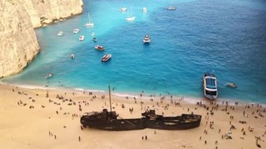 Yunanistan 'ın Zakynthos kentindeki İyon Denizi kıyısındaki Navagio plajının insansız hava aracı görüntüsü. Demir atmış tekneler, dinlenen insanlar, paslı gemi, mavi su.
