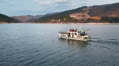 Romanya 'daki Bicaz gölünde yüzen insanlarla dolu bir teknenin insansız hava aracı görüntüsü. Tepeler yemyeşil ormanlarla kaplı.