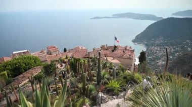 MENTON, FRANCE - Eylül, 2021: tropikal bitkilerle park. İnsanlar, geleneksel konut binalarının çatıları, arka planda deniz.