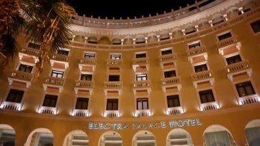 SALONIKI, GREECE - EYLÜL, 2020: Gece lambaları, yürüyen insanlar ve park halindeki arabalarla merkez meydandaki Electra Palace Hotel