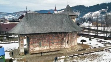 Romanya 'daki Mizah Manastırı' nın kışın insansız hava aracı görüntüsü. Ana boyanmış kilise ve kule, karlı ve turistli avlu, çıplak ağaçlar, konut binaları