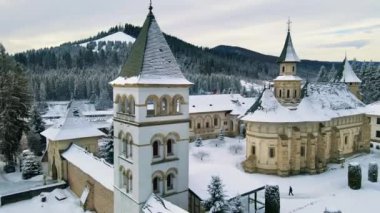 Romanya 'daki Putna Manastırı' nın kışın insansız hava aracı görüntüsü. Yürüyen turistlerle, çıplak ağaçlarla, karla, tepelerle dolu bir bahçe.