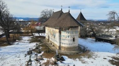 Romanya 'daki Arbore Manastırı' nın kışın insansız hava aracı görüntüsü. Ana boyanmış kilise, karlı ve mezarlıklı bahçe, çıplak ağaçlar, konut binaları.