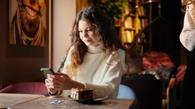 Kafedeki kahveciye Uluslararası Aşı Sertifikası COVID-19 ile akıllı telefon gösteren bir kadın. Arttırıcı doz. QR kodunu tarıyorum. Kafenin içinde, içinde pasta olan bir masa.