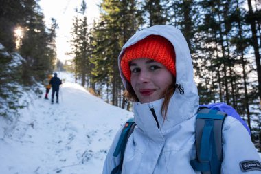 Beyaz kayak kostümü ve parlak turuncu şapkası olan beyaz, beyaz, beyaz bir kadın yürüyüşçünün portresi. Kış mevsimi. Yol karla kaplı ve öndeki diğer yürüyüşçüler.