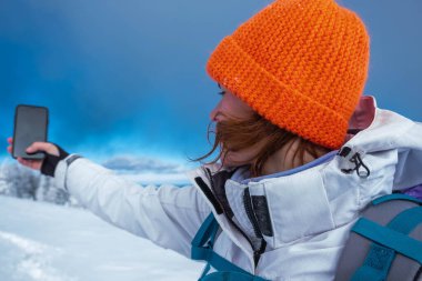 Beyaz kayak kostümü ve parlak turuncu şapkasıyla fotoğraf çeken beyaz, beyaz, beyaz bir kadın yürüyüşçünün portresi. Kış mevsimi. Yürüyüş karla kaplıydı ve diğer yürüyüşçüler öndeydi.