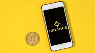 CHISINAU, MOLDOVA - Mart 2022: Fiziksel Bitcoin altın madeni para, Binance uygulaması bir telefonda açıldı, sarı arkaplan