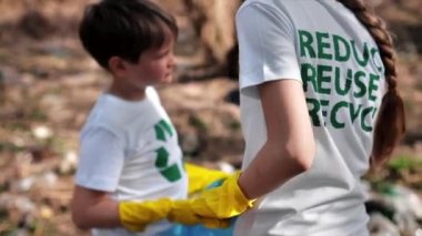 Erkek ve kız, kirli bir temizleme yerindeki torba içinde plastik çöp topluyor tişörtlerdeki geri dönüşüm işaretlerini topluyor. Yavaş çekim