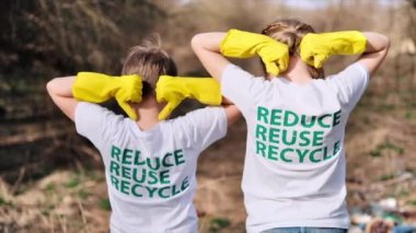 Lastik eldivenli kız ve erkek tişörtlerindeki ekolojik yazıları gösteriyor. Kirli bir açıklıkta plastik çöp toplama