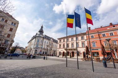 CLUJ, ROMANIA - Nisan 2022: Şehir merkezinin sokak manzarası. Yürüyen insanlar, arabalar, eski binalar ve bir anıt.