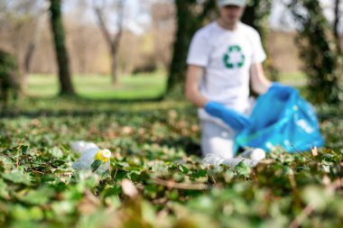 Kirli bir parkta plastik çöp toplayan bir adam. Lastik eldivenler, tişörtte geri dönüşüm tabelası.