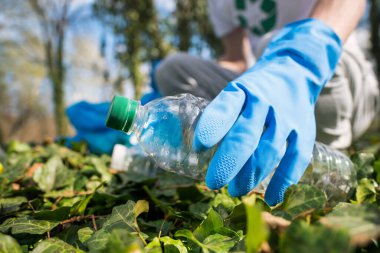 Kirli bir parktaki konteynırda plastik çöp toplayan bir adam. Lastik eldivenler, tişörtte geri dönüşüm işaretleri.