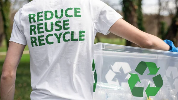 Man at plastic garbage collecting in a polluted park. Rubber gloves, holding a container with plastic bottles. Recycling signs on the T-shirt and container