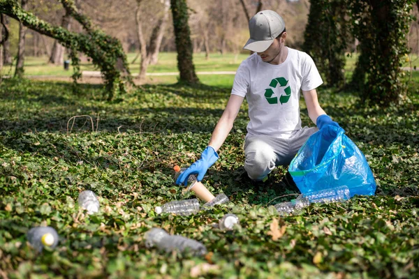Man collecting plastic garbage in bag in a polluted park. Rubber gloves, recycling sign on T-shirt