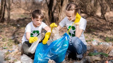 Erkek ve kız, kirlenmiş bir temizleme yerindeki torba içinde plastik çöp topluyor tişörtlerdeki geri dönüşüm işaretleri...