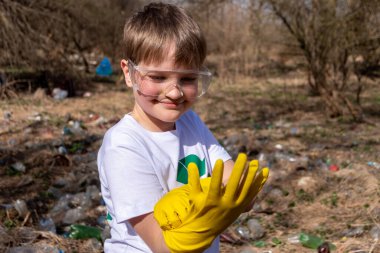 Tişörtünde geri dönüşüm sembolü ve gözlükleriyle çevredeki plastiğin ön planına sarı eldivenler takan beyaz beyaz bir çocuk.