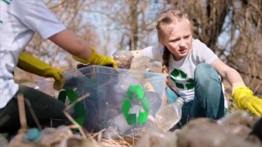Erkek ve kız, kirlenmiş bir temizleme kabında plastik çöp topluyor tişörtlerdeki geri dönüşüm tabelalarını topluyor. Yavaş çekim
