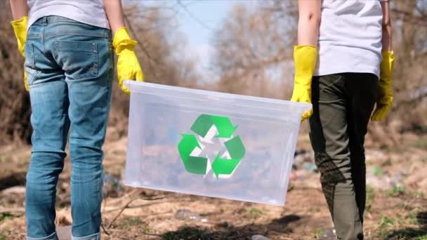 穿着橡胶手套的男孩和女孩拿着一个集装箱 在一个被污染的地方收集塑料垃圾 回收T恤衫上的标识 慢动作 — 图库视频影像