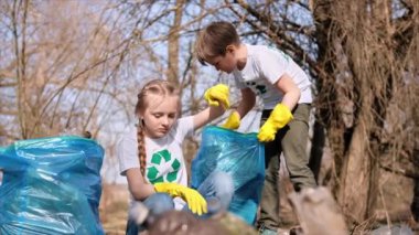 Erkek ve kız, kirli bir temizleme yerindeki torba içinde plastik çöp topluyor tişörtlerdeki geri dönüşüm işaretlerini topluyor. Yavaş çekim