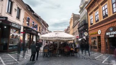 BRASOV, ROMANIA - Nisan 2022: Eski şehir merkezinin zaman gösterimi. Bir sürü yürüyen insan, restoran ve eski binalarla dolu bir yaya sokağı.