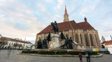 CLUJ, ROMANIA - Nisan 2022: Şehir merkezinin gün batımında sokak manzarası, zaman aşımı. Aziz Michael Kilisesi ile merkez meydanı ve önünde Matthias Corvinus heykeli, yürüyen ve dinlenen insanlar