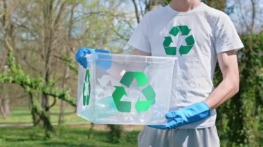 Kirli bir parkta çöpleri toplayan bir adam. Plastik eldivenler, plastik şişelerle dolu bir konteynır. Tişört ve konteynırdaki geri dönüşüm işaretleri