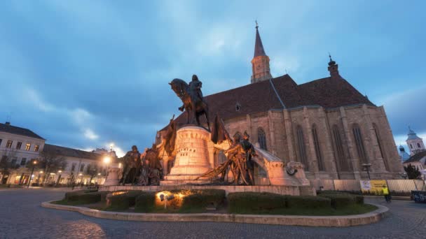 Cluj Romania エイプリル 2022 夕方の都心の街並み タイムラプス 聖ミヒャエル教会とその前にマティアス コルヴィヌスの像と中央広場 歩行と休憩の人々 — ストック動画