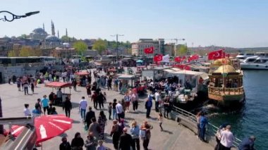 İSTANBUL, TURKEY - Nisan 2022: Şehir merkezindeki dolgu caddesi. Bir sürü yürüyen ve dinlenen insan, demirlemiş tekneler, binalar, arabalar.