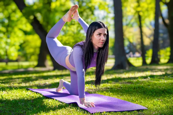 Attractive girl with long black hair dressed in purple doing yoga pose - One leg pigeon, in the green park