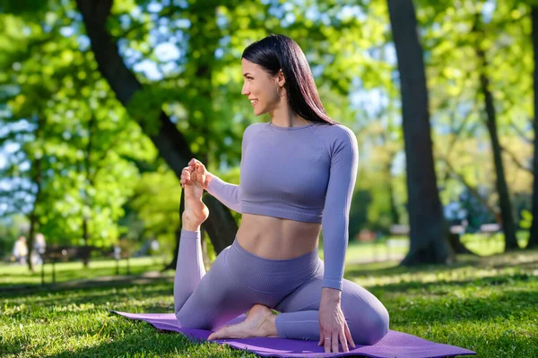 Attractive girl with long black hair dressed in purple doing yoga pose in the green park