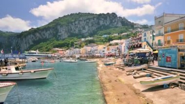CAPRI, İtalya - Mayıs 2022: kasabanın deniz limanının görüntüsü. Bağlı tekneler, binaları ve yürüyen insanları olan toprak seti caddesi, yeşillik.