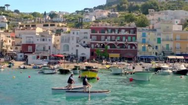 CAPRI, İtalya - Mayıs 2022: kasabanın deniz limanının görüntüsü. Bağlı tekneler, binaları ve yürüyen insanları olan toprak seti caddesi, yeşillik.
