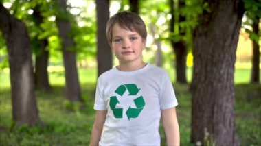 Plastik eldivenli bir çocuk kirli bir parktaki plastik çöp toplama yerinde tişörtünde geri dönüşüm işareti varmış gibi görünüyor. Yavaş çekim