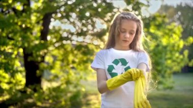 Kirli bir parkta plastik çöp toplamak için lastik eldiven takan bir kız tişörtte geri dönüşüm tabelası takıyor. Yavaş çekim