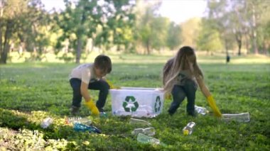 Erkek ve kız, kirli bir parktaki konteynırda plastik çöp topluyor tişörtlerdeki geri dönüşüm işaretlerini topluyor. Yavaş çekim