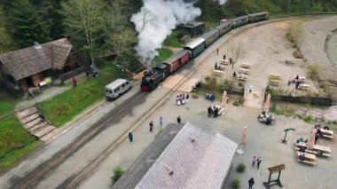 VISEU DE SUS, ROMANIA - MAYIS, 2022: Mocanita buharlı treninin demiryolu istasyonundaki bir vadideki hava aracı görüntüsü, dinlenen turistler, yemyeşil orman ve nehir çevresi