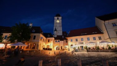 SIBIU, ROMANIA - MAYIS 2022: akşam kentin Tarih Merkezi 'nin zaman gösterimi. Kare şeklinde kule ve önünde insanlar, klasik tarzda yapılmış eski binalar.