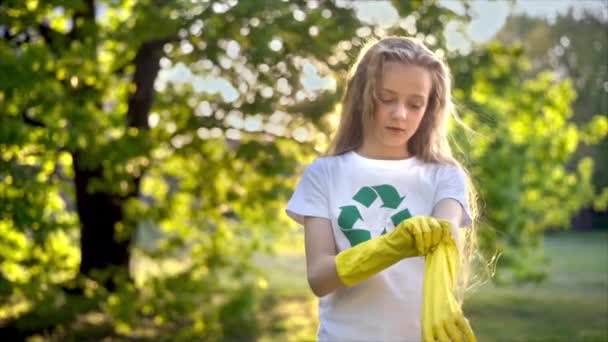 女孩戴上橡胶手套 在被污染的公园里收集塑料垃圾 T恤上的回收标志 慢动作 — 图库视频影像