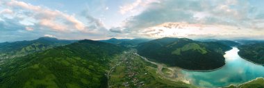 Romanya 'daki Karpatlar' da hava aracı doğaya panoramik açıdan bakıyor. Izvorul Gölü Muntelui ve vadide bir köy, yemyeşil ormanlarla kaplı tepeler.