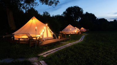 Tents with burning torches, lamps and wooden chairs at glamping, forest around, dusk