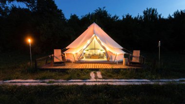 Tent interior and exterior with burning torches, lamps and wooden chairs at glamping, forest around, dusk