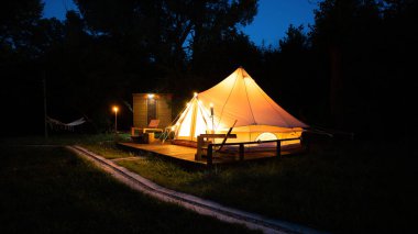 Tent with burning torches, lamps and wooden chairs at glamping, forest around, dusk